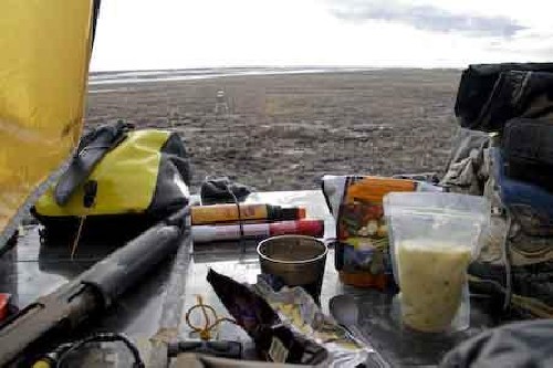 View out through tent vestibule. Note the dehydrated food, mug of hot chocolate, flare, bear spray, cocolate wrapper, shotgun, and bear alarm tripwire tripod in distance.